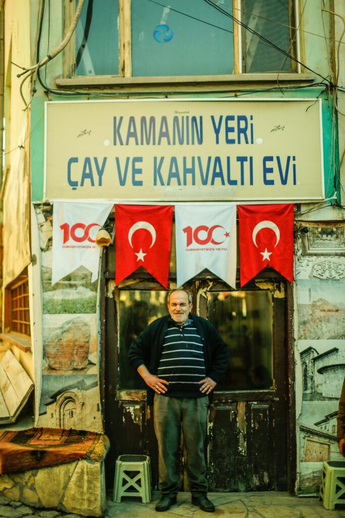 Man posing in front of a traditional Turkish cafe adorned with flags and signage.